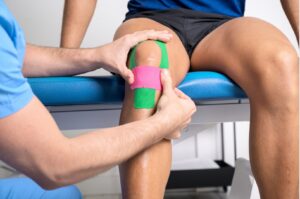 Physiotherapeut klebt Kinesio-Tape auf das Knie eines Patienten.